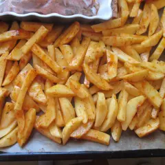 Gotowe do pieczenia frytki w piekarniku, pokrojone ziemniaki doprawione przyprawami, leżą na blasze wyłożonej papierem.