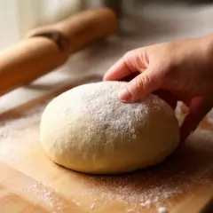 Dłoń dotyka kulki ciasta na pizzę posypanej mąką na desce, obok wałek.