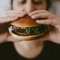 Osoba trzymająca w dłoniach cheeseburgera z podwójnym kotletem i zieloną sałatą.