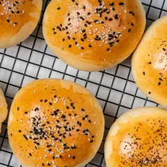 Złociste bułeczki do mini burgerów posypane sezamem i czarnuszką, na czarnej kratce.