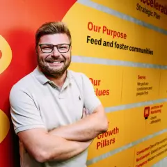 Uśmiechnięty mężczyzna w okularach na tle ściany z hasłami McDonald's. Zastanawiasz się, ile jest McDonaldów w Polsce? Ta ściana może pomóc w rozwoju sieci.