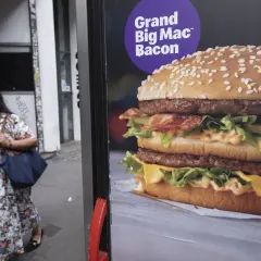 Reklama Grand Big Mac Bacon. Kobieta w kwiecistej sukience mija billboard z apetycznym burgerem.