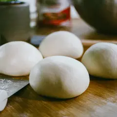 Cztery kulki surowego ciasta na pizzę leżą na drewnianym blacie obok noża.