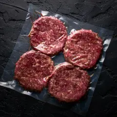 Cztery surowe kotlety wołowe, gotowe do przyprawienia mięsa na hamburgery, zapakowane próżniowo na czarnym tle.