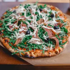 Apetyczna pizza prosciutto z rukolą, oliwkami i parmezanem, podana na papierze.