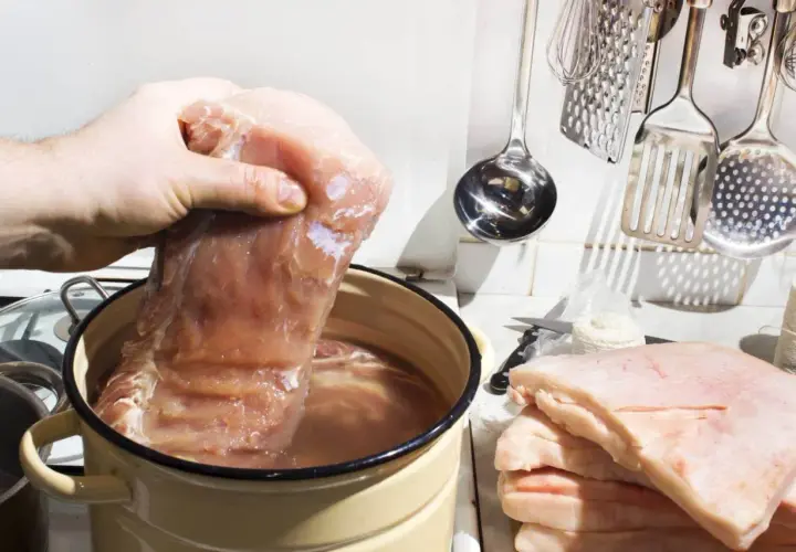 Ręka zanurza kawałek mięsa w garnku z wodą, przygotowując solankę do mięsa. Ile soli na litr wody? Obok leży stos słoniny.