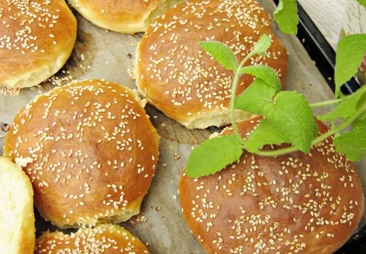 Złociste bułki do burgerów posypane sezamem, ułożone na papierze do pieczenia, z gałązką mięty.
