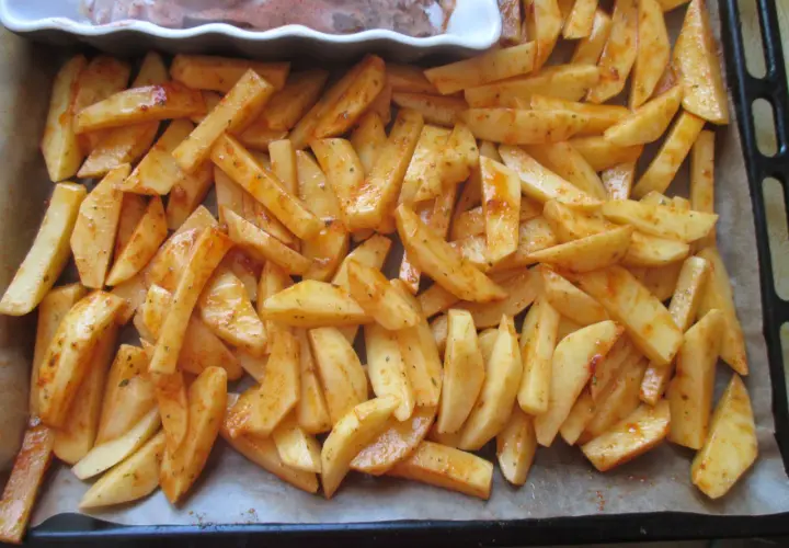 Gotowe do pieczenia frytki w piekarniku, pokrojone ziemniaki doprawione przyprawami, leżą na blasze wyłożonej papierem.
