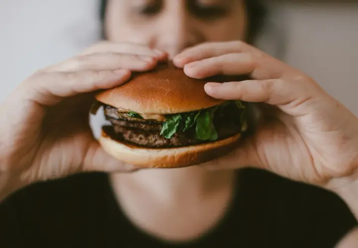 Osoba trzymająca w dłoniach cheeseburgera z podwójnym kotletem i zieloną sałatą.