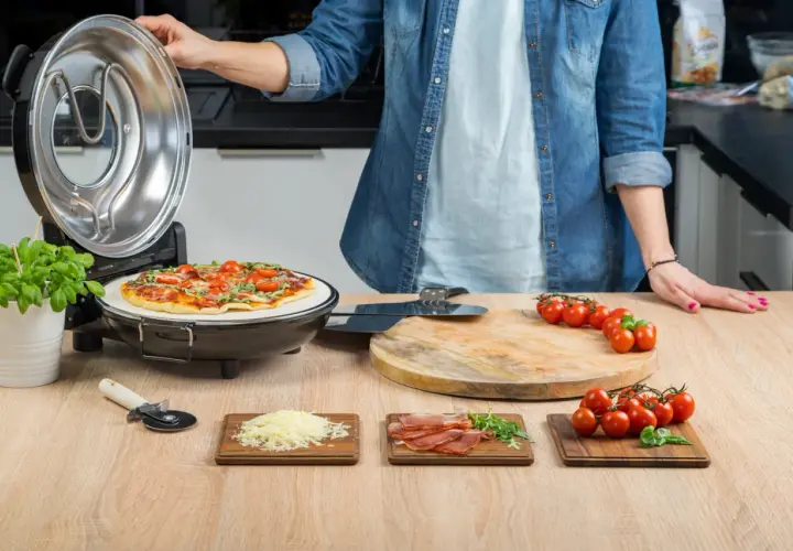 Osoba zamyka pokrywę pieca do pizzy, obok gotowa pizza i składniki.