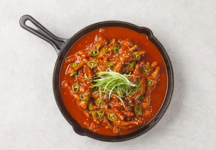Kurczak w sosie gochujang, podany w żeliwnej patelni, ozdobiony zieloną cebulką i sezamem.