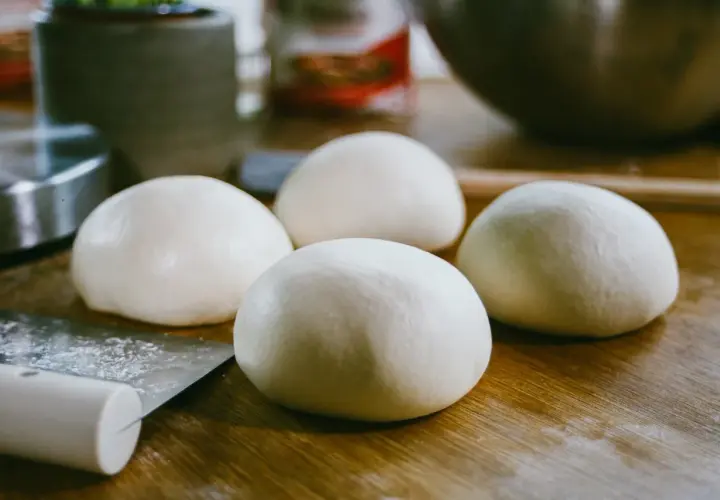 Cztery kulki surowego ciasta na pizzę leżą na drewnianym blacie obok noża.