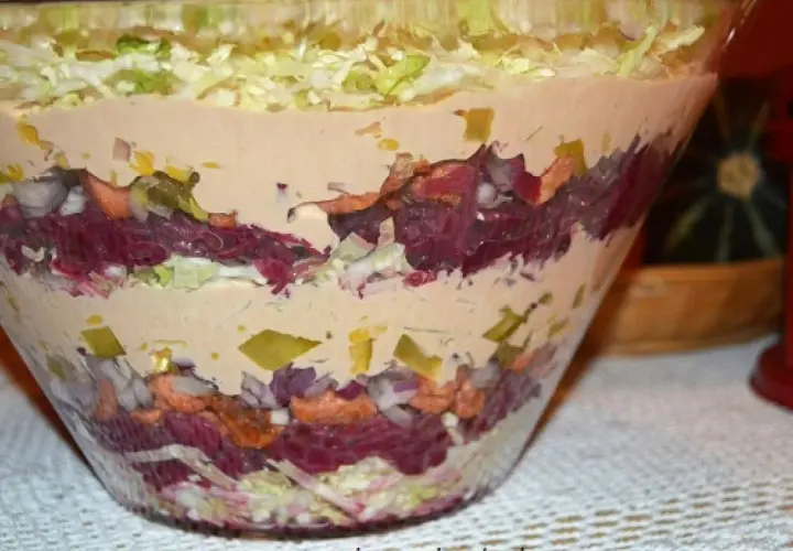 Kolorowa sałatka warstwowa, przypominająca gyros warstwy, w szklanej misie. Widoczne warstwy kapusty, buraków, cebuli i sosu.