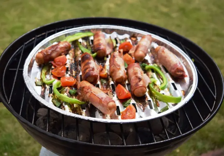 Pyszna kiełbasa na grilla, owinięta boczkiem, pieczona z papryką i pomidorkami na aluminiowej tacce.