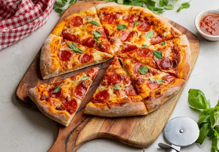 Apetyczna pizza pepperoni na drewnianej desce, ozdobiona listkami bazylii. Obok sos pomidorowy i czerwono-biała serwetka.
