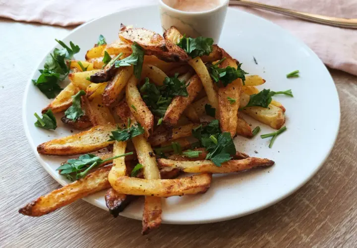 Złociste frytki z natką pietruszki na białym talerzu, obok sosu.