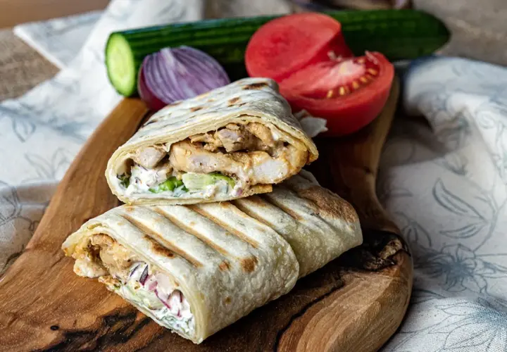 Pyszny kebab w tortilli z kurczakiem, warzywami i sosem, podany na drewnianej desce z pomidorem i ogórkiem.
