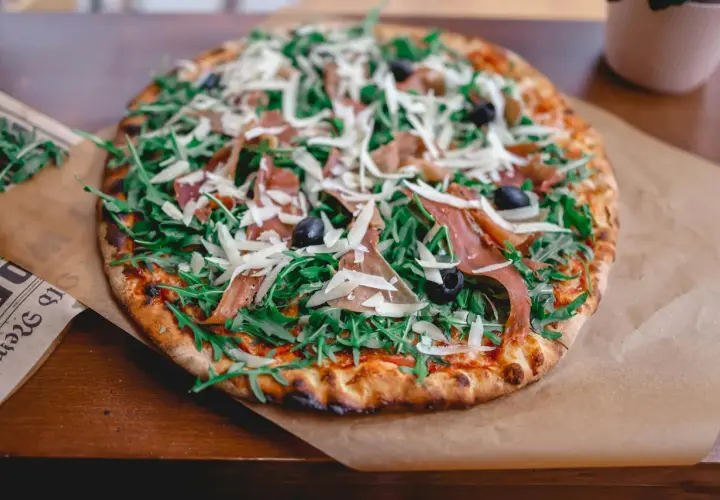 Apetyczna pizza prosciutto z rukolą, oliwkami i parmezanem, podana na papierze.
