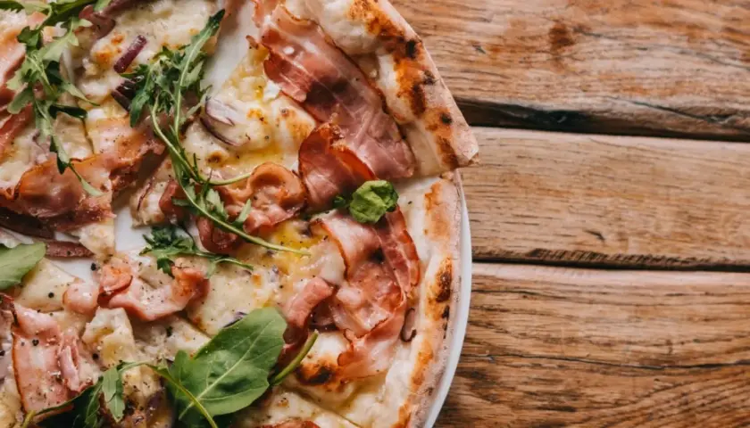 Pyszna pizza carbonara z chrupiącym boczkiem, rukolą i cebulą na drewnianym blacie.