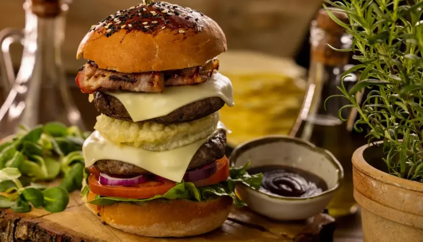 Wielowarstwowy burger wołowy z bekonem, serem, warzywami i sosem na drewnianej desce.