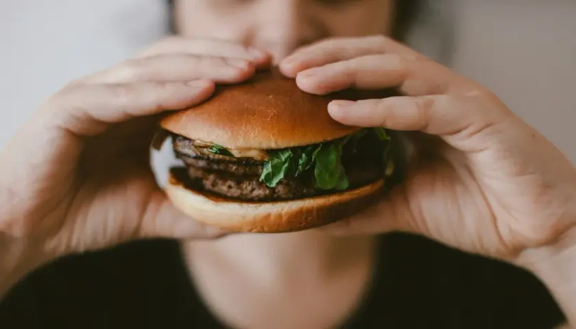 Osoba trzymająca w dłoniach cheeseburgera z podwójnym kotletem i zieloną sałatą.
