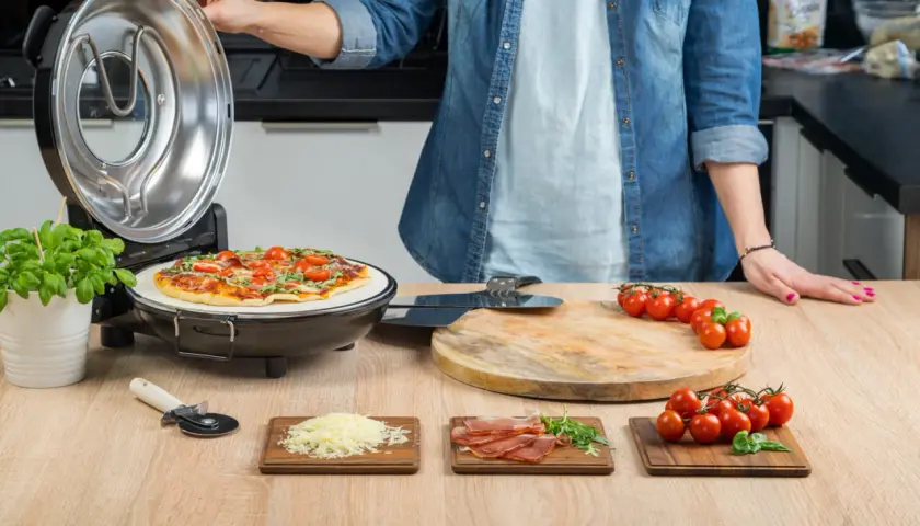 Osoba zamyka pokrywę pieca do pizzy, obok gotowa pizza i składniki.
