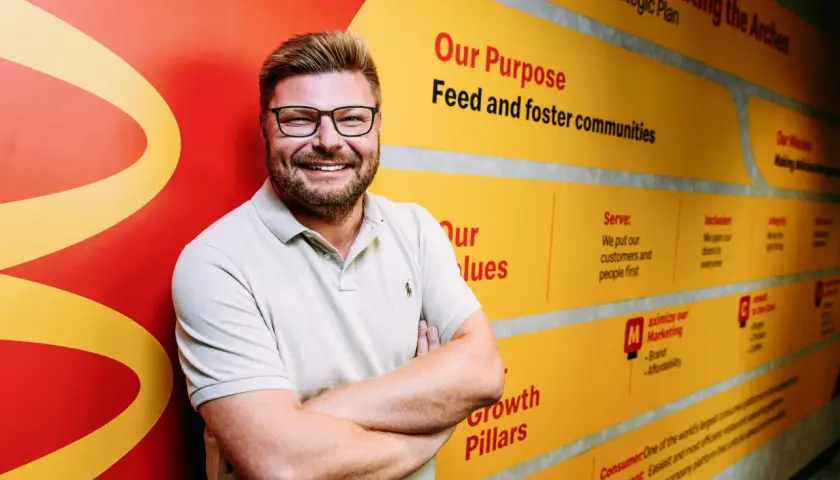 Uśmiechnięty mężczyzna w okularach na tle ściany z hasłami McDonald's. Zastanawiasz się, ile jest McDonaldów w Polsce? Ta ściana może pomóc w rozwoju sieci.