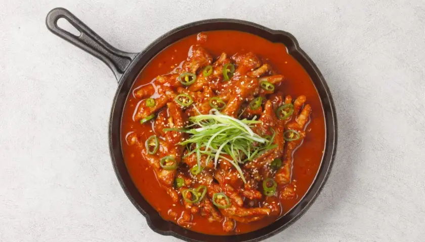 Kurczak w sosie gochujang, podany w żeliwnej patelni, ozdobiony zieloną cebulką i sezamem.