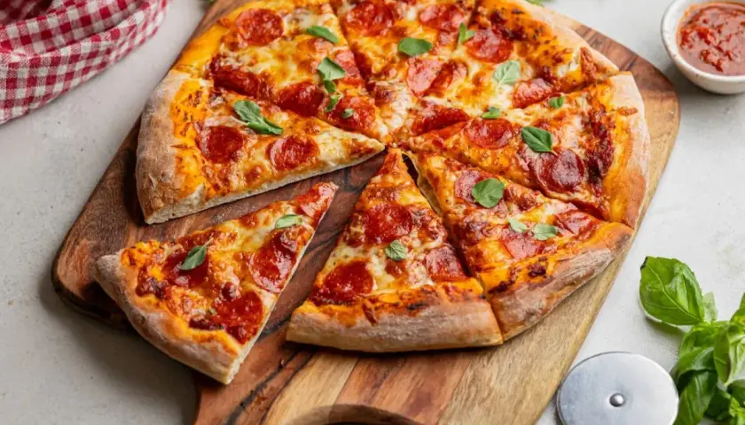 Apetyczna pizza pepperoni na drewnianej desce, ozdobiona listkami bazylii. Obok sos pomidorowy i czerwono-biała serwetka.