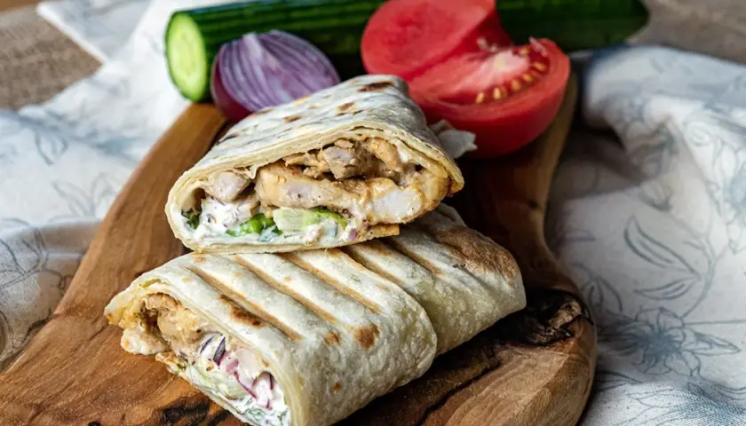 Pyszny kebab w tortilli z kurczakiem, warzywami i sosem, podany na drewnianej desce z pomidorem i ogórkiem.