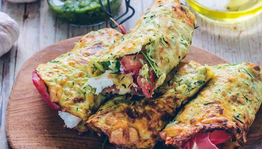 Złociste tortilla z cukinii, nadziewane serem, pomidorami i szynką, prezentowane na drewnianej desce.