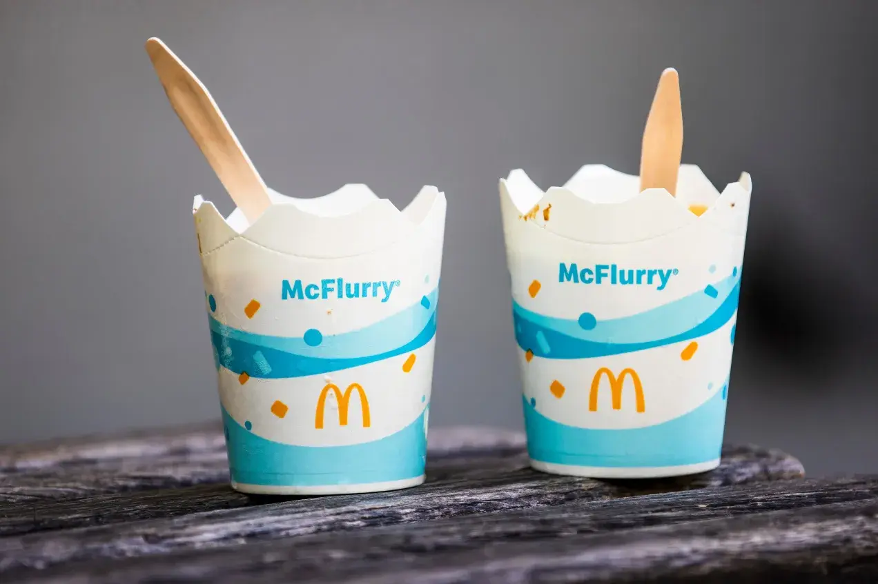 Dwa kubeczki McFlurry pistacjowe z drewnianymi łyżeczkami na drewnianym blacie.