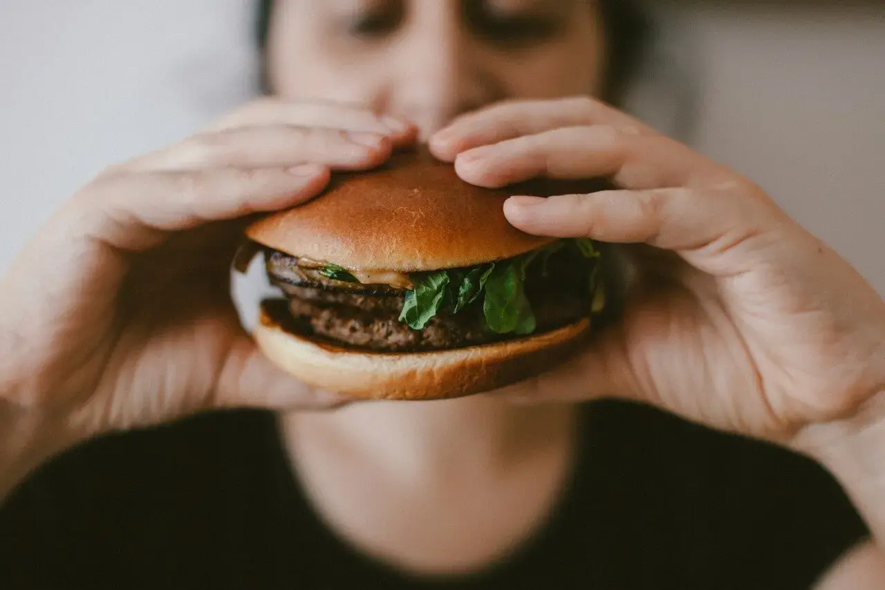 Osoba trzymająca w dłoniach cheeseburgera z podwójnym kotletem i zieloną sałatą.