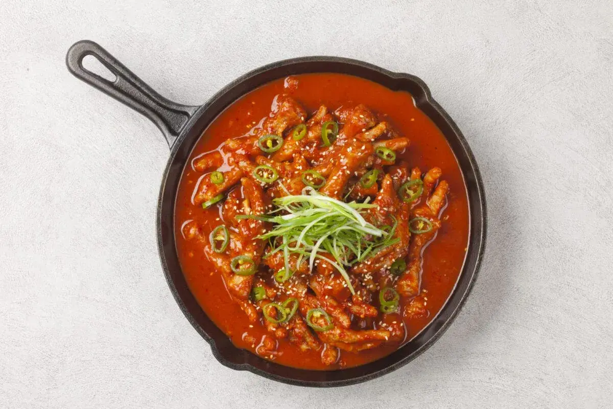 Kurczak w sosie gochujang, podany w żeliwnej patelni, ozdobiony zieloną cebulką i sezamem.