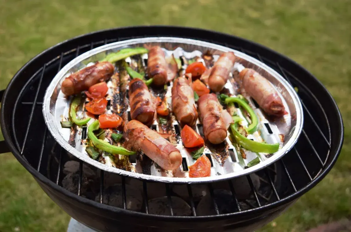 Pyszna kiełbasa na grilla, owinięta boczkiem, pieczona z papryką i pomidorkami na aluminiowej tacce.