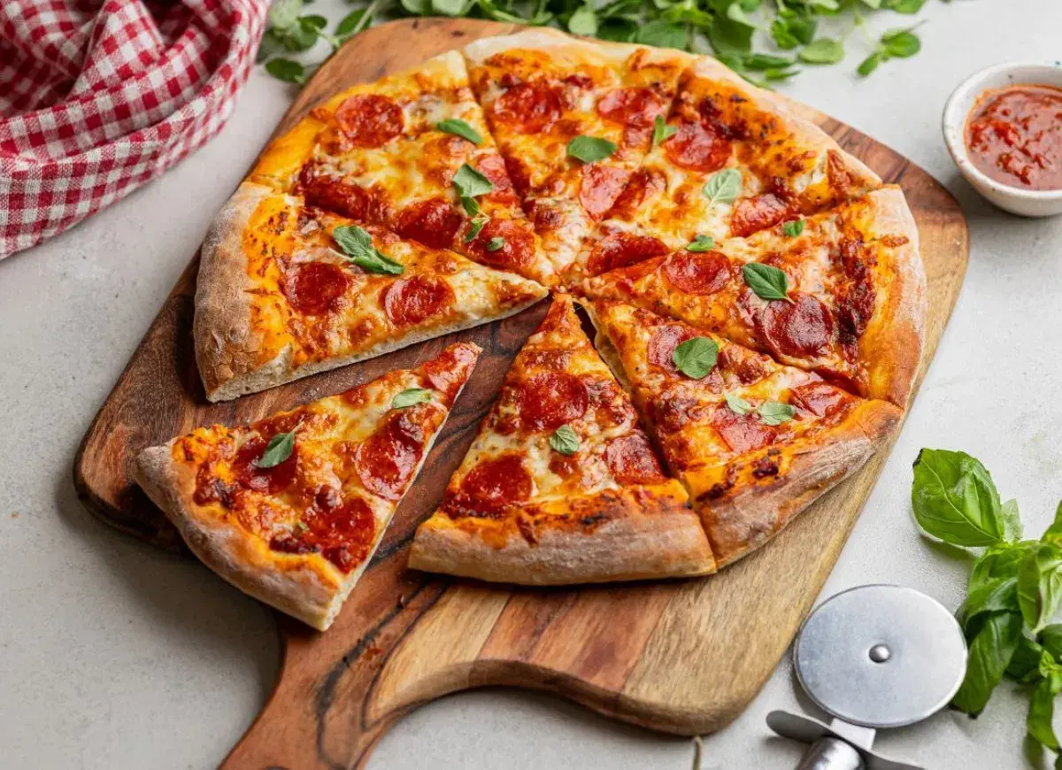 Apetyczna pizza pepperoni na drewnianej desce, ozdobiona listkami bazylii. Obok sos pomidorowy i czerwono-biała serwetka.