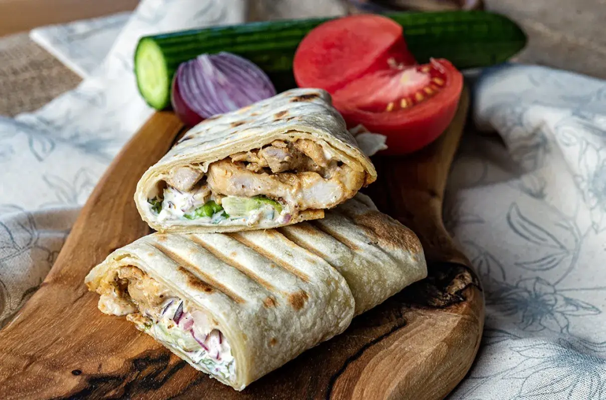 Pyszny kebab w tortilli z kurczakiem, warzywami i sosem, podany na drewnianej desce z pomidorem i ogórkiem.