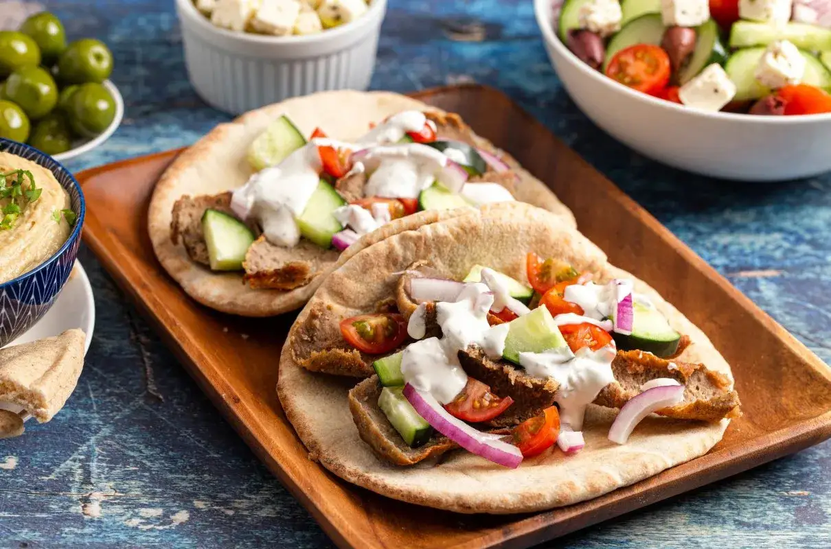 Pyszne gyros składniki: mięso, pomidory, ogórek, cebula i sos na chlebku pita. Obok miseczka z hummusem i oliwkami.