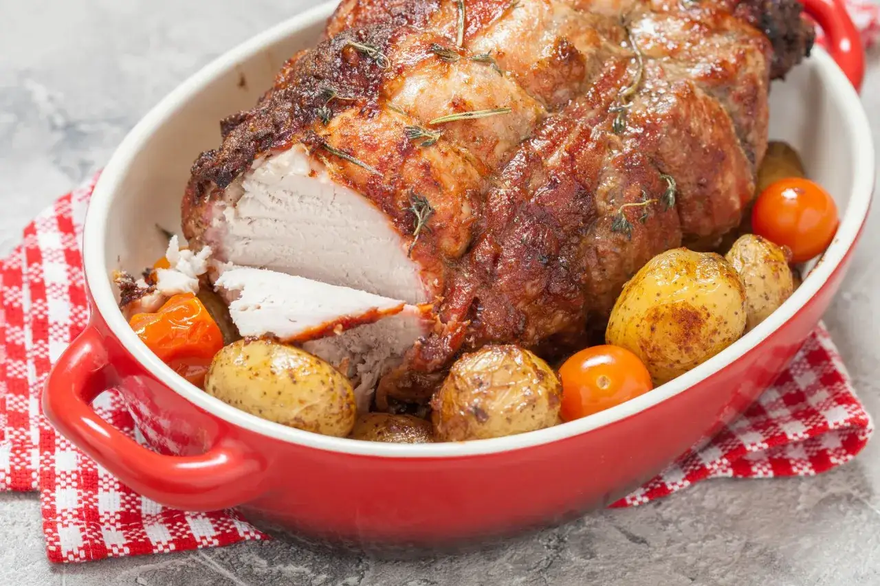 Pieczony schab w czerwonym naczyniu z ziemniakami i pomidorkami, idealny po marynacie.