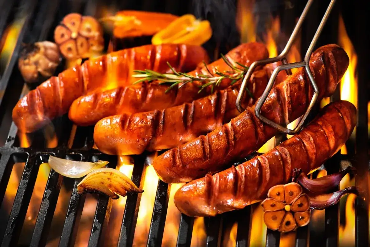 Soczysta kiełbasa na grilla skwierczy na ogniu, obok pieczony czosnek i cebula. Idealna na letnie wieczory.