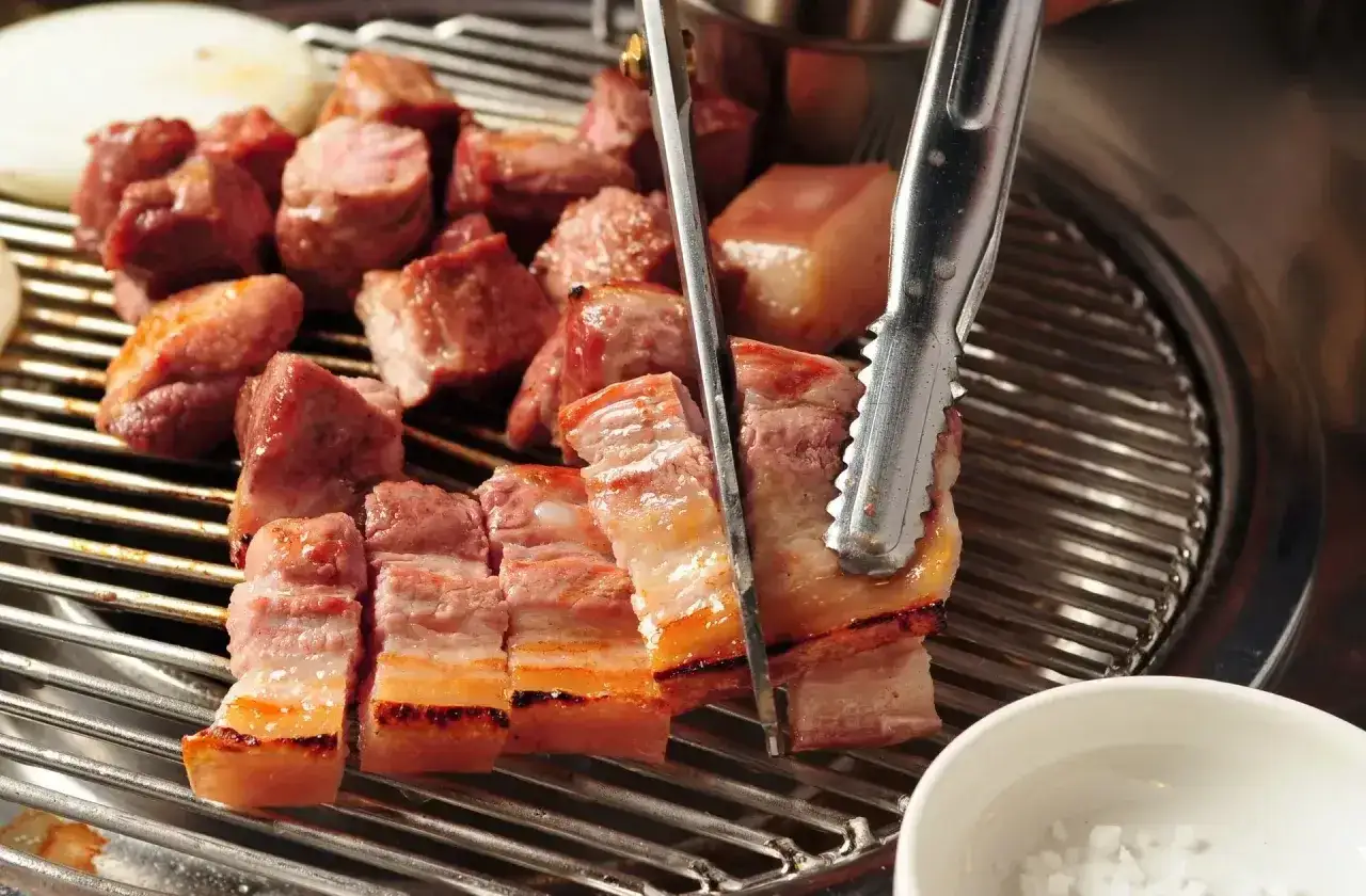 Soczysty boczek na grillu, idealny do marynaty. Kawałki mięsa pieką się na ruszcie, a szczypce obracają je, by równomiernie się zarumieniły.