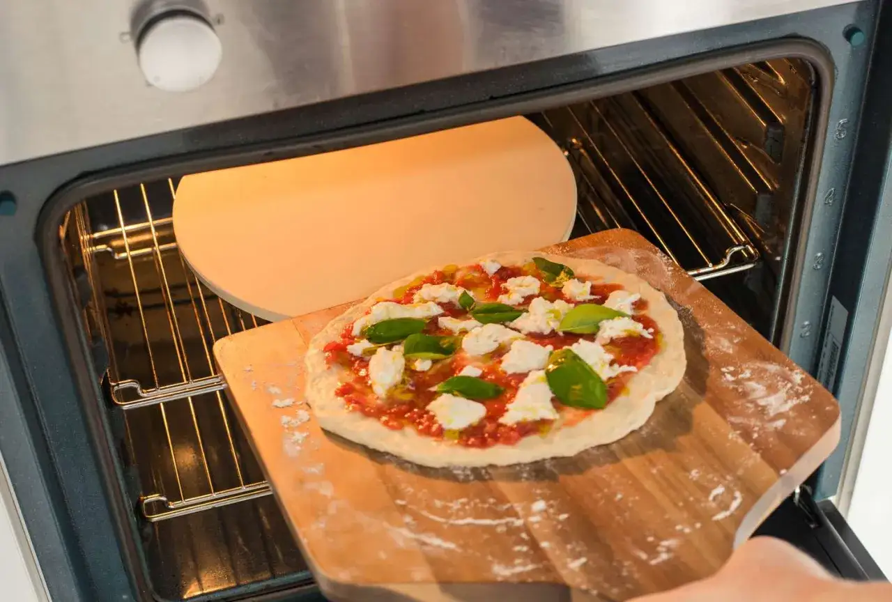Przygotowanie pizzy na dzień pizzy 2026. Surowe ciasto z sosem, mozzarellą i bazylią na desce, wsuwane do rozgrzanego piekarnika.