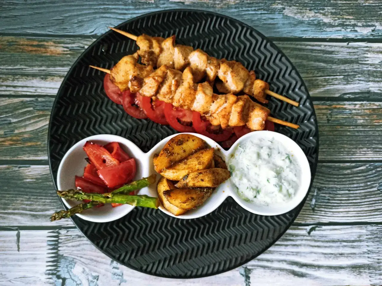 Dwa szaszłyki z kurczaka, czyli pyszne souvlaki, podane z pieczonymi ziemniakami, pieczoną papryką, szparagami i sosem tzatziki.