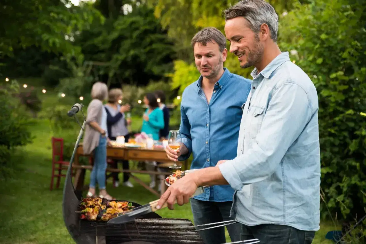 Dwóch mężczyzn przy grillu, przygotowuje nowości na grilla. W tle widać gości i girlandę światełek.